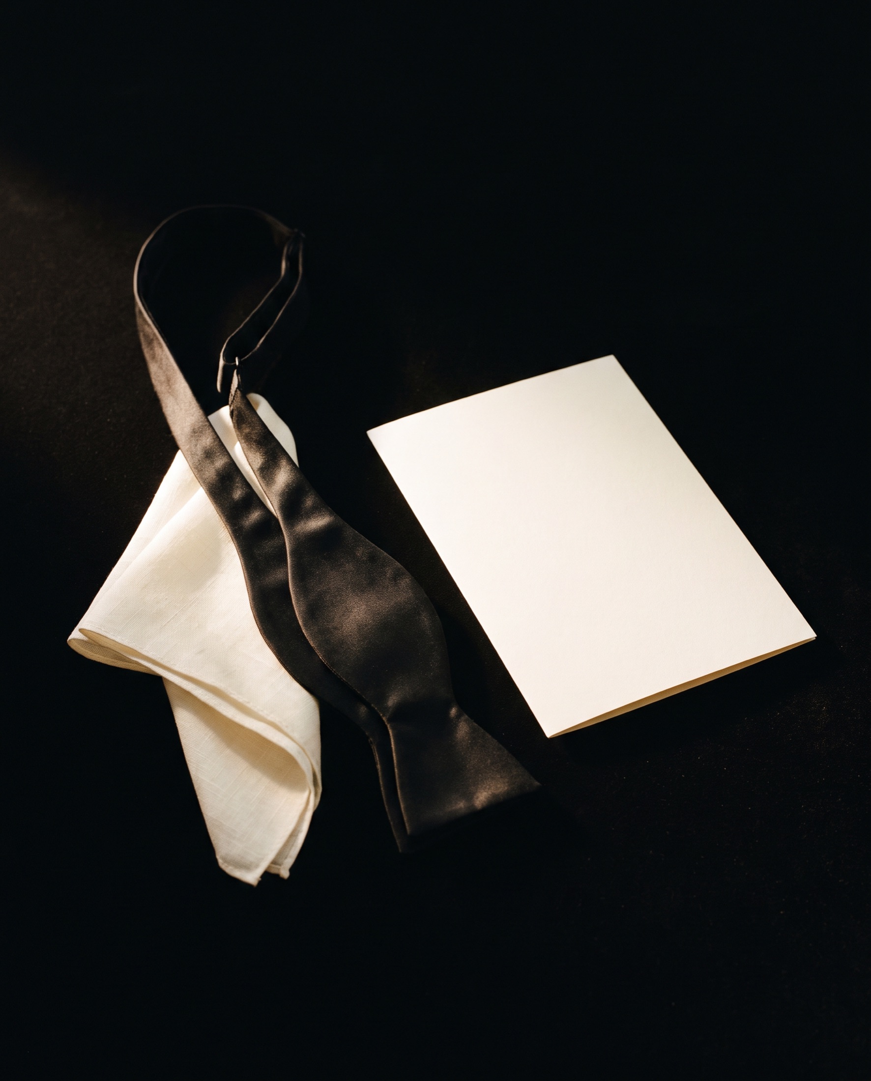 A black silk bow tie, an ivory pocket square and a blank invitation card on black velvet